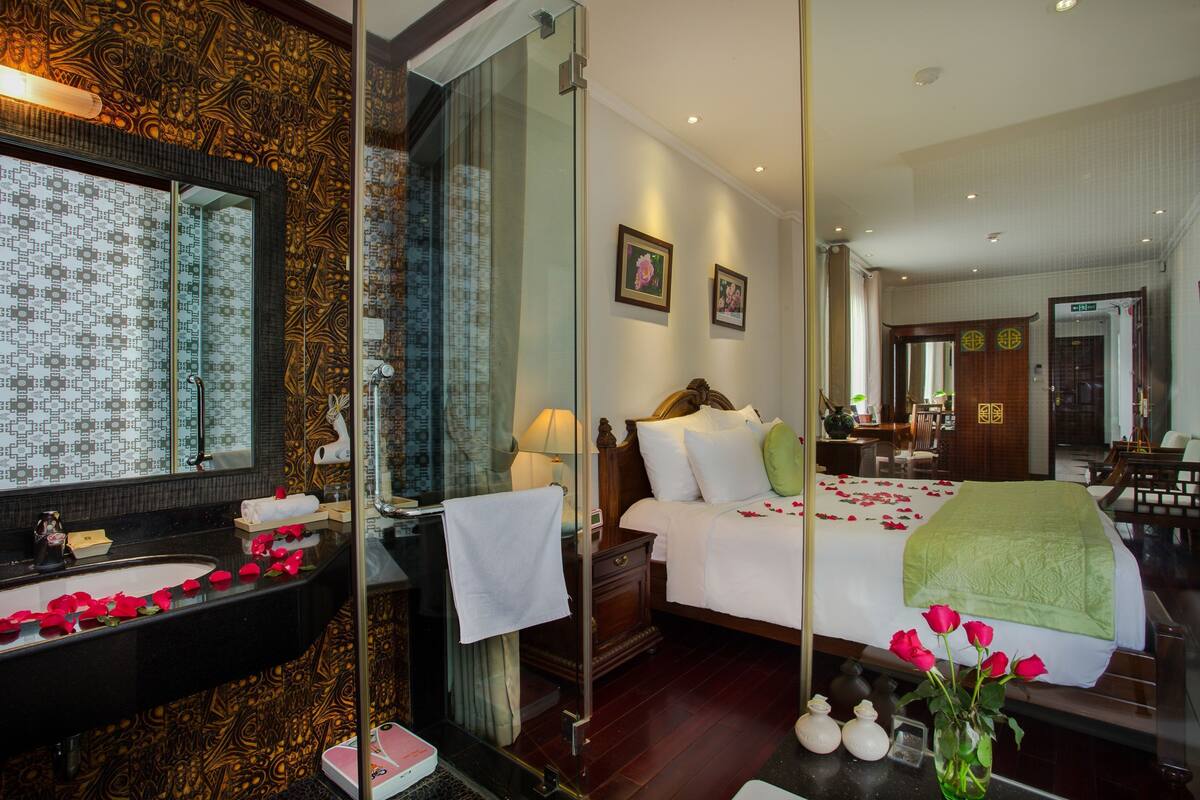 A hotel room with a large mirror, a bed with rose petals, a bedside table with a vase of flowers, and a bathroom with a patterned sink.