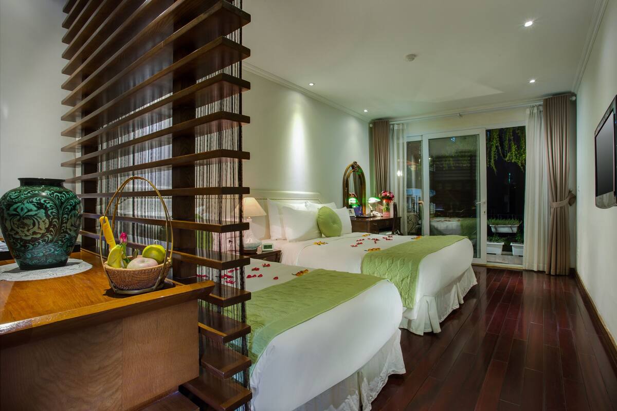 A hotel room with a wooden staircase, two beds with green and white bedding, a bedside table with a lamp, a vase, and a basket with fruits.