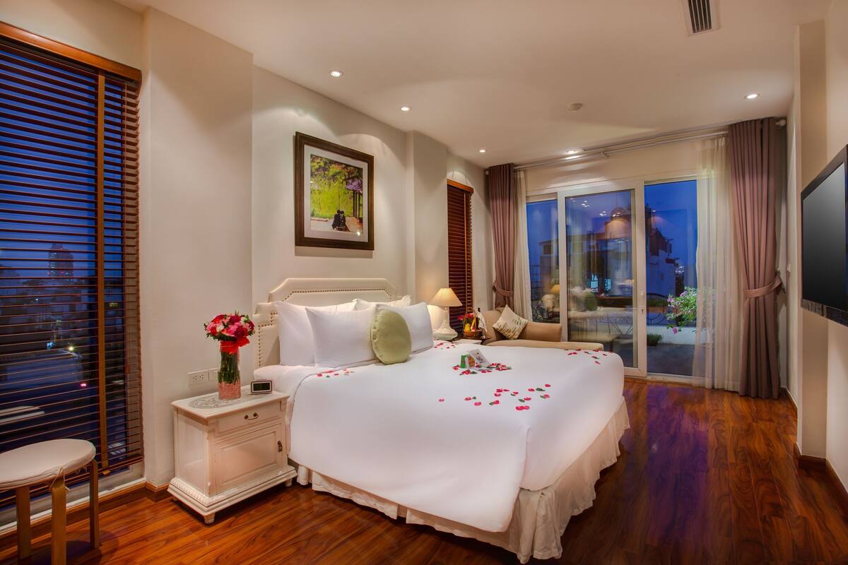 A hotel room with a large bed, a nightstand, a chair, a window with blinds, and a view of the outside.