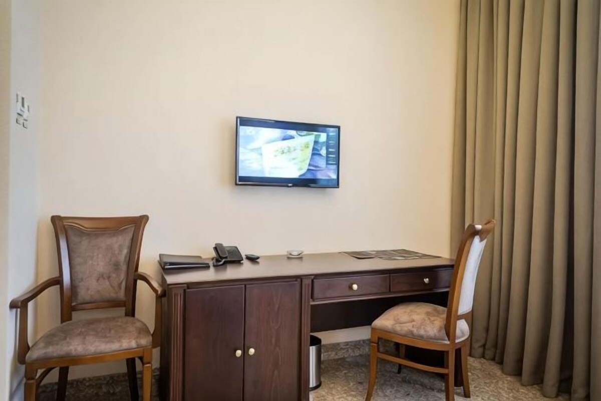 A hotel room with a desk, two chairs, a television mounted on the wall, and a window with curtains.