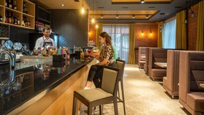 Zona de bar con un camarero, un cliente sentado y diversas botellas y vasos en estanterías.