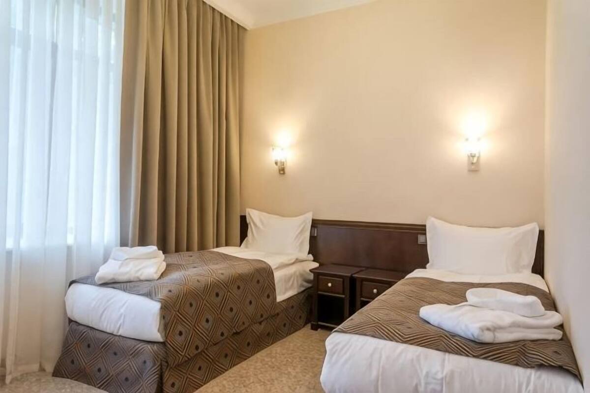 A hotel room with two beds, a nightstand, a lamp, and a window with curtains.
