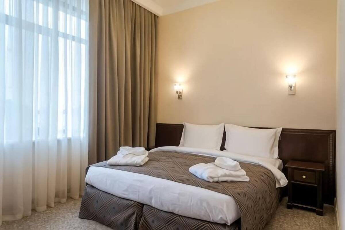 A hotel room with a large bed, two bedside tables, a nightstand, and a window with curtains.