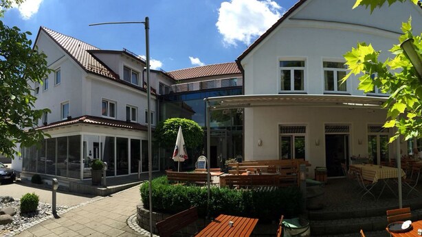 Un patio con mobiliario exterior, una zona cubierta y un edificio con fachada blanca y tejado rojo.