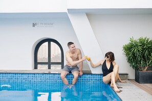 Ein Mann und eine Frau sitzen an einem Pool mit einer gefliesten Kante, einem Topfpflanze und einem Fenster mit Blick auf Gebäude.
