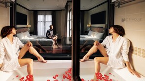Una stanza con specchi, un letto, una persona seduta sul bordo di una vasca e petali di rosa sparsi sul pavimento.
