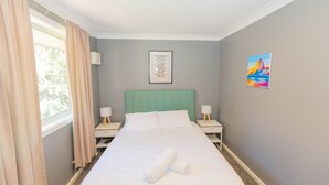 Ein ordentlich bezogenes Bett mit weißen Leinen und einem aufgerollten Handtuch, ein grün gepolsterter Kopfteil, zwei Nachttische mit Lampen, eine gerahmte Liste mit Winteraktivitäten an der Wand und ein farbenfrohes Gemälde an der rechten Wand.