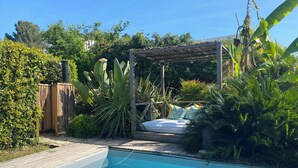 Une piscine avec une terrasse en bois, un espace salon couvert et une végétation luxuriante.