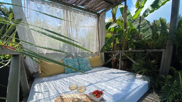 Un lit avec un drap blanc, deux oreillers, un plateau en bois avec des verres et un bol de fruits, et une terrasse en bois.