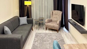 A modern living room with a grey sofa, a beige armchair, a small coffee table, a TV mounted on the wall, and a ceiling light fixture. - Beta Homes Boutique Hotel (Izmir)