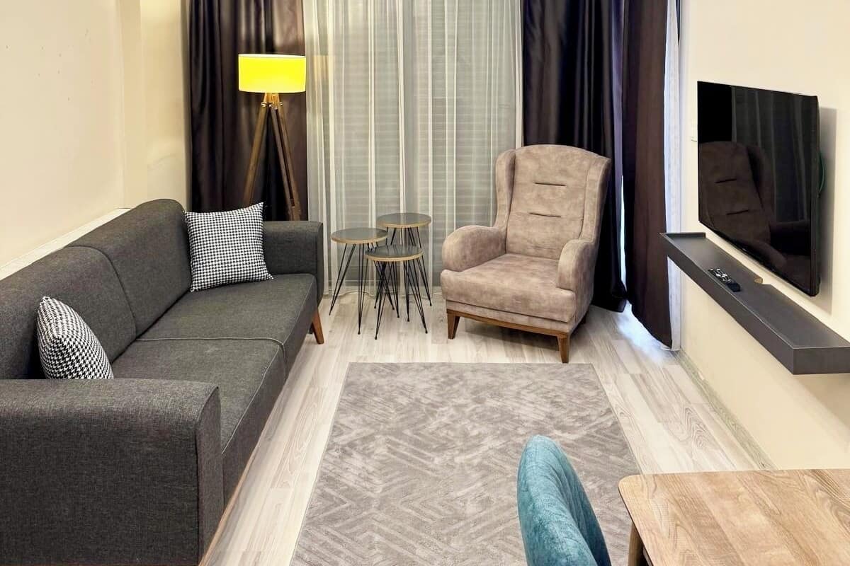 Una sala de estar moderna con un sofá gris, un sillón beige, una mesita de centro, un televisor montado en la pared y una lámpara en el techo.