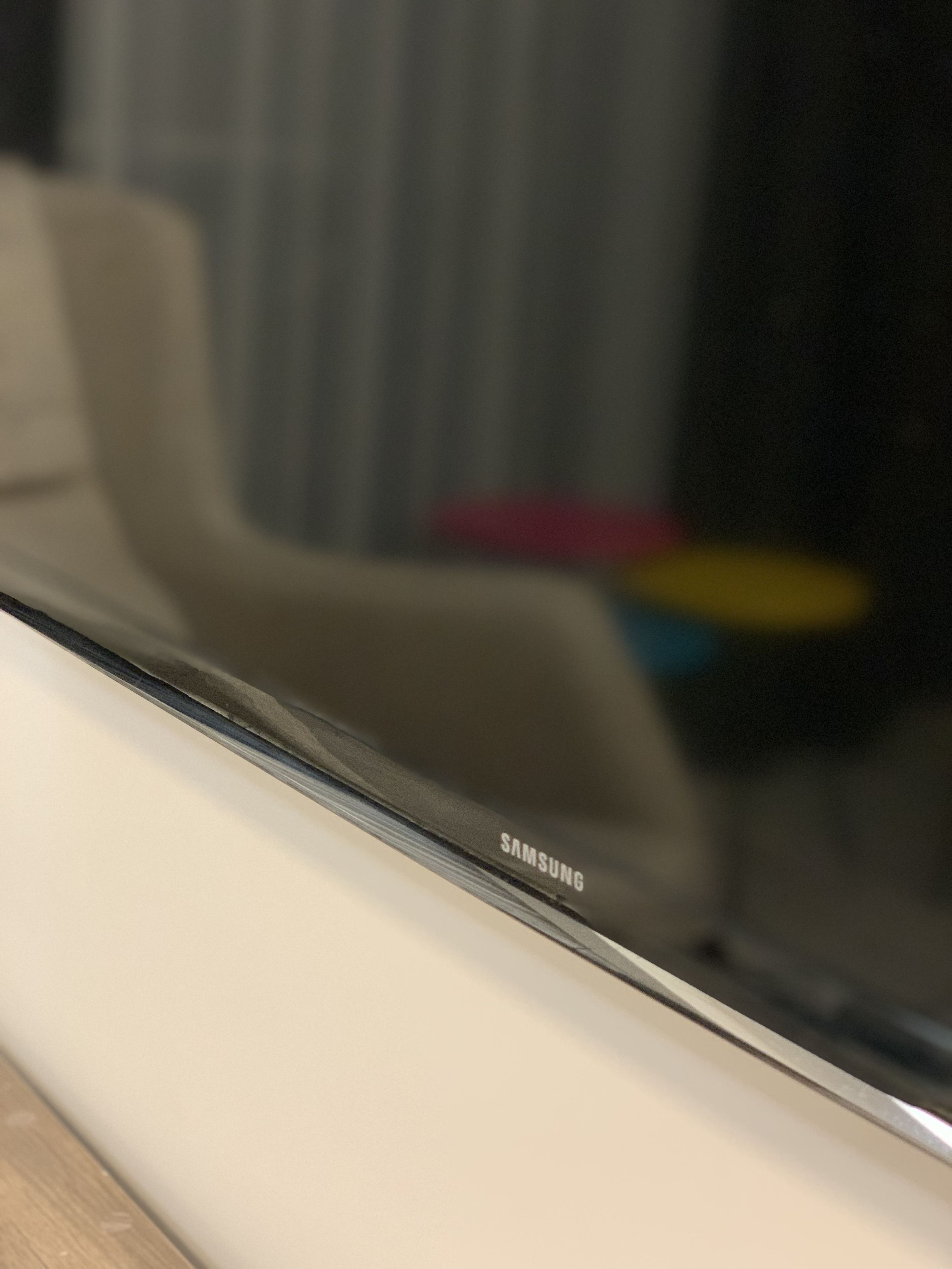 Primer plano de un dispositivo Samsung, posiblemente un teléfono inteligente, con un fondo desenfocado que incluye una silla y un objeto de colores.