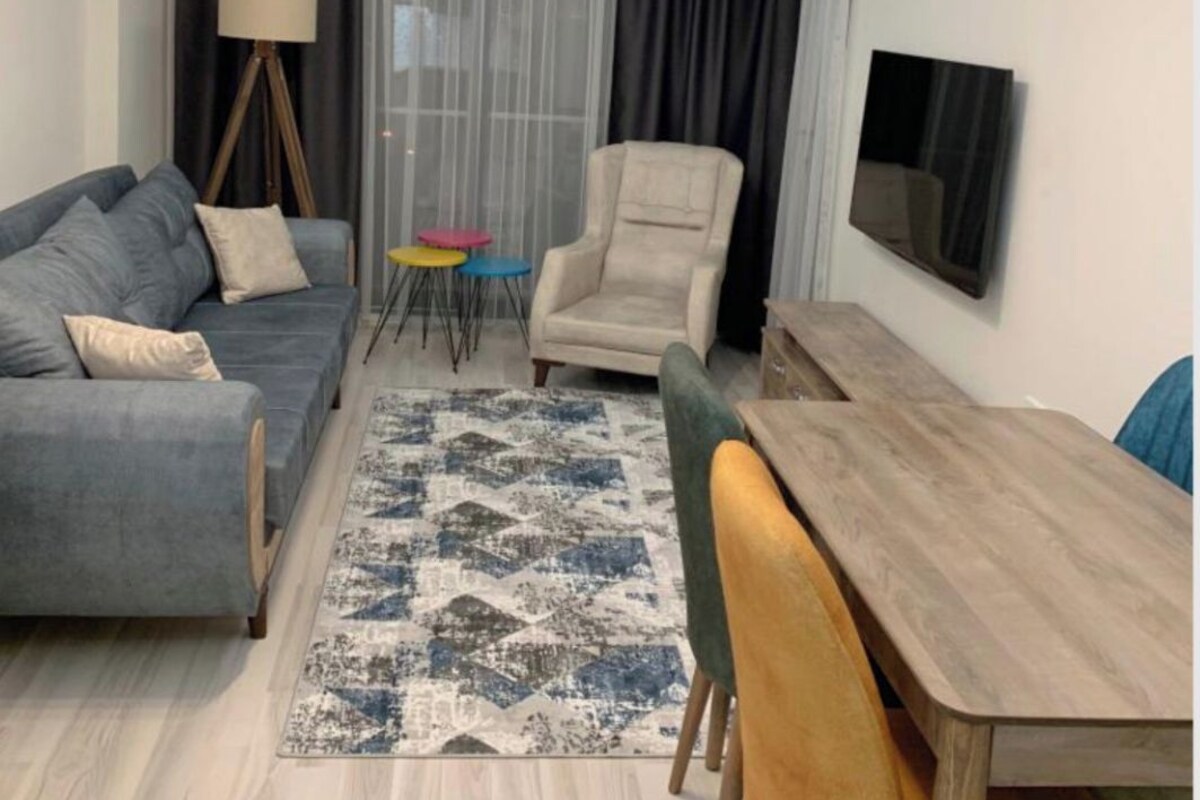 A modern living room with a sofa, armchair, dining table, and a TV mounted on the wall.