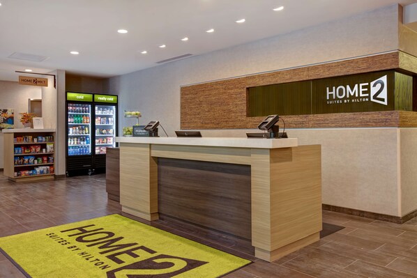 A hotel reception area with a reception desk, a minibar, a refrigerator stocked with beverages, and a small kitchenette. - Home2 Suites by Hilton Charlotte Northlake (Charlotte)