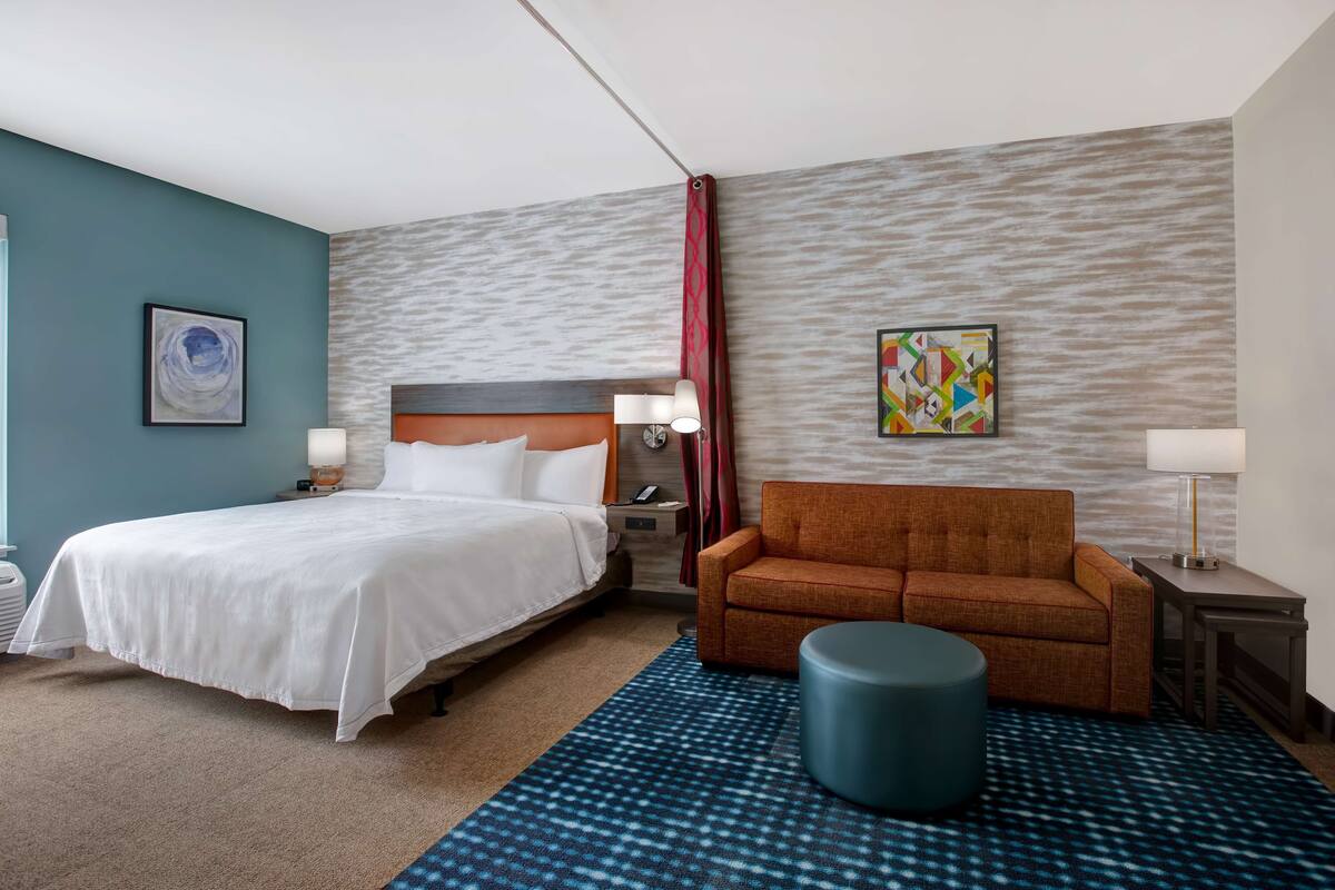 Ein Hotelzimmer mit Bett, Sofa, runder Hocker, Nachttisch, Lampe und zwei Wandbildern.