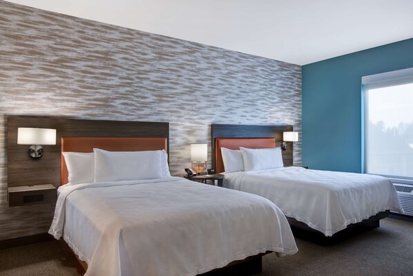 A hotel room with two beds, each with white bedding and headboards, a nightstand with a lamp, and a large window with blinds.