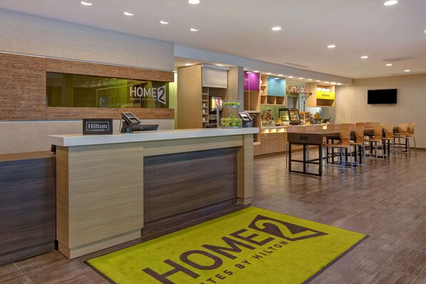 Zona de recepción de un hotel con mostrador, un cartel de 'Home 2 Suites by Hilton', una estación de café y un área de descanso con sillas.