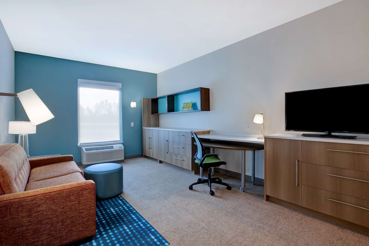 Ein modernes Hotelzimmer mit Sofa, Schreibtisch, Fernseher und Fenster mit Blick auf Bäume.