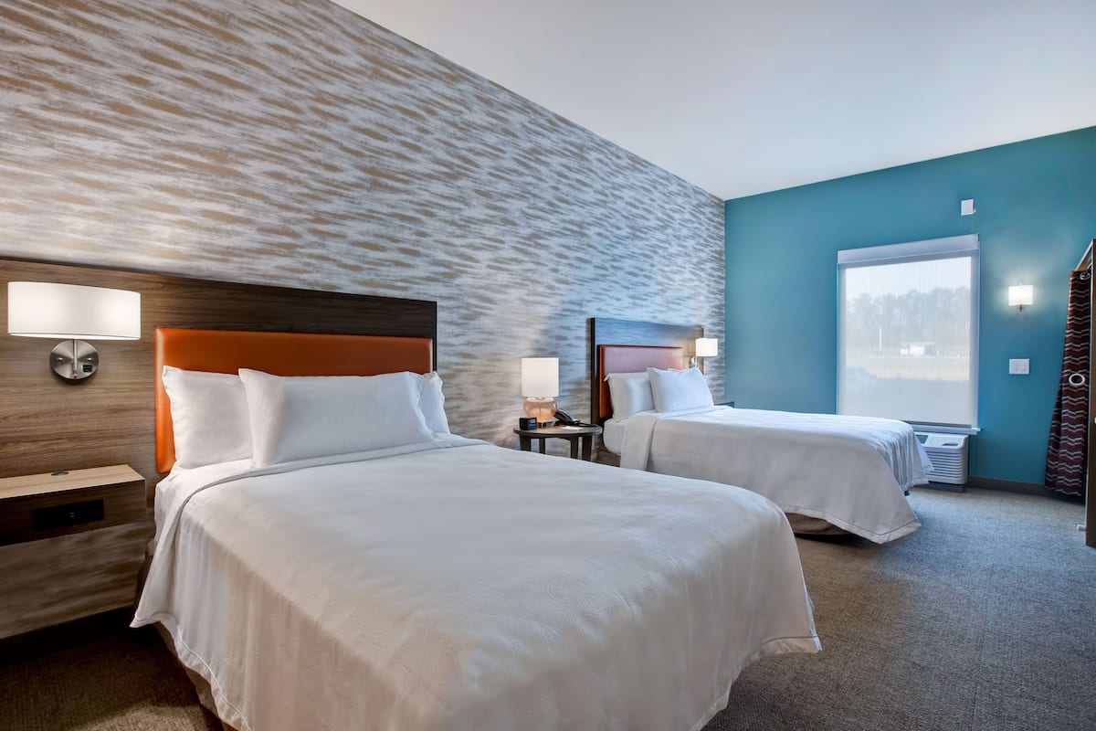 Ein Hotelzimmer mit zwei Betten, einem hölzernen Kopfteil, einer Wand mit strukturierter Oberfläche, einem Fenster mit Jalousien und einem kleinen Tisch mit einer Lampe.