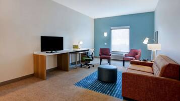 A modern hotel room with a desk, television, two chairs, a sofa, and a large window.