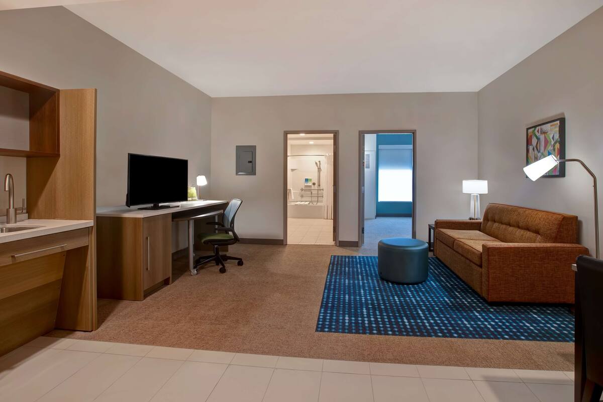 Ein modernes Hotelzimmer mit Küchenzeile, einem Flachbildfernseher, einer Couch, einem Couchtisch und einem durch eine offene Tür einsehbaren Badezimmer.