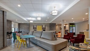 A modern lounge area with a sofa, chairs, a table, and a refrigerator. - Home2 Suites by Hilton Charlotte Northlake (Charlotte)