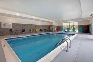 Una piscina interior con un cartel de advertencia sobre la ausencia de socorrista, sillas de descanso de varios colores y una vista despejada del exterior a través de amplias ventanas.