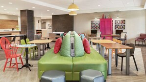 A modern lounge area with a green sofa, multiple tables, and chairs, and a reception desk in the background. - Home2 Suites by Hilton Harrisburg North (Harrisburg)