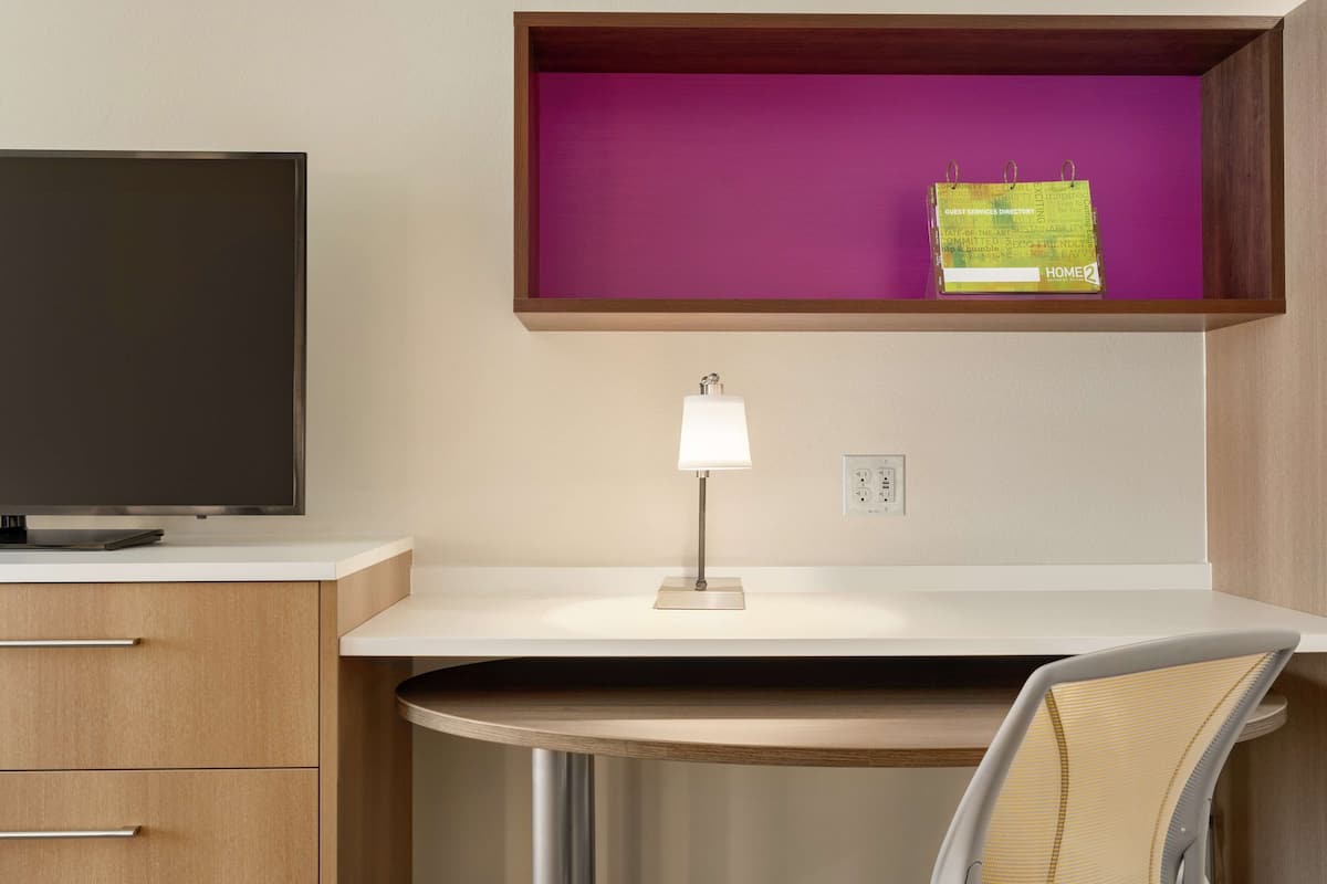 Une chambre d’hôtel moderne équipée d’une télévision à écran plat, d’un bureau avec une lampe, d’une chaise et d’une étagère violette contenant un répertoire des services à la disposition des clients.
