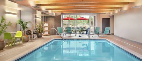 A swimming pool with a wooden ceiling, green chairs, and red umbrellas.