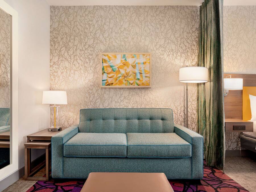 A hotel room with a teal sofa, a small table with a lamp, a painting on the wall, and a bed with a headboard.