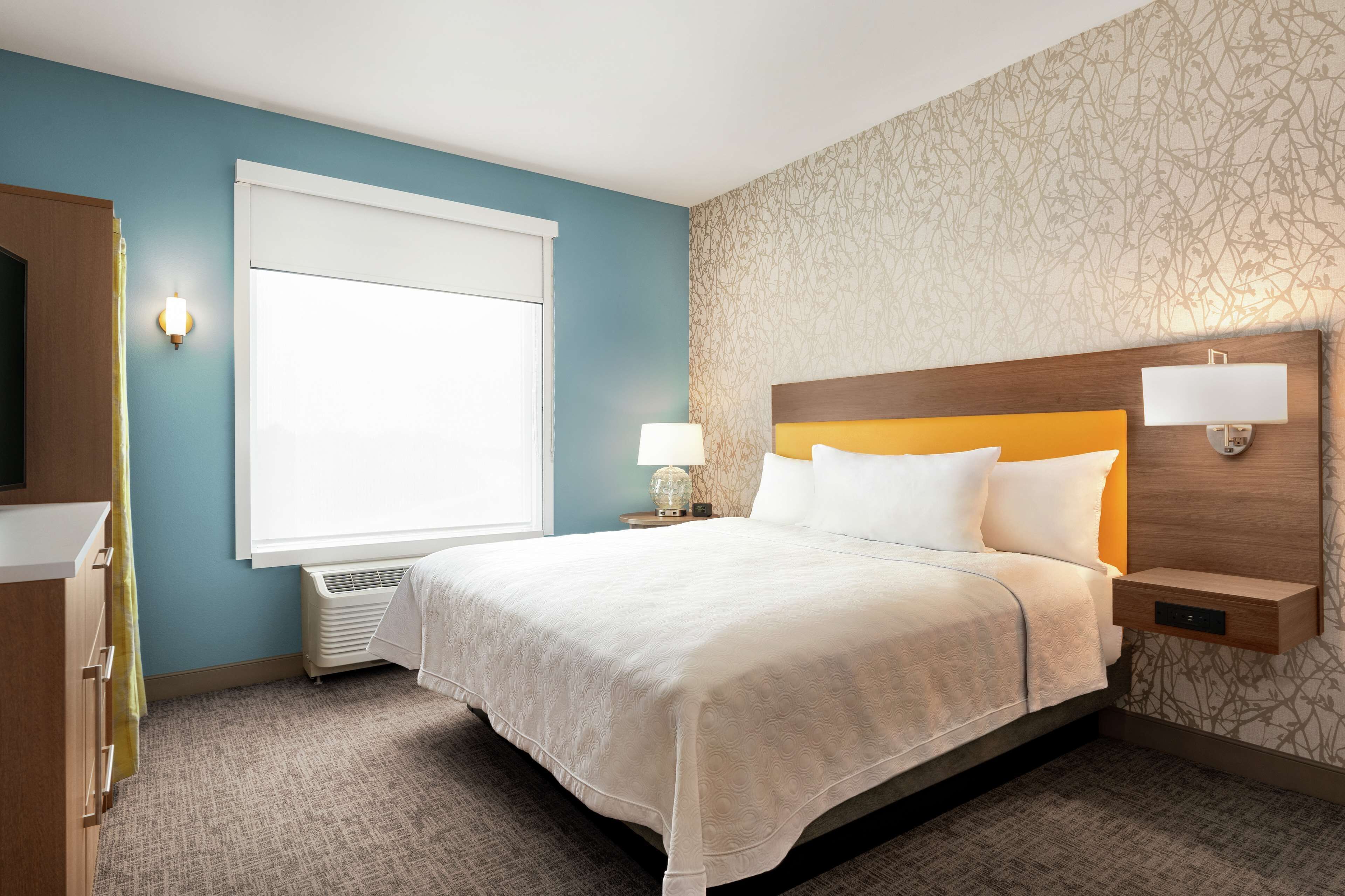 A hotel room with a large bed, a bedside table with a lamp, a wardrobe, and a window with blinds.