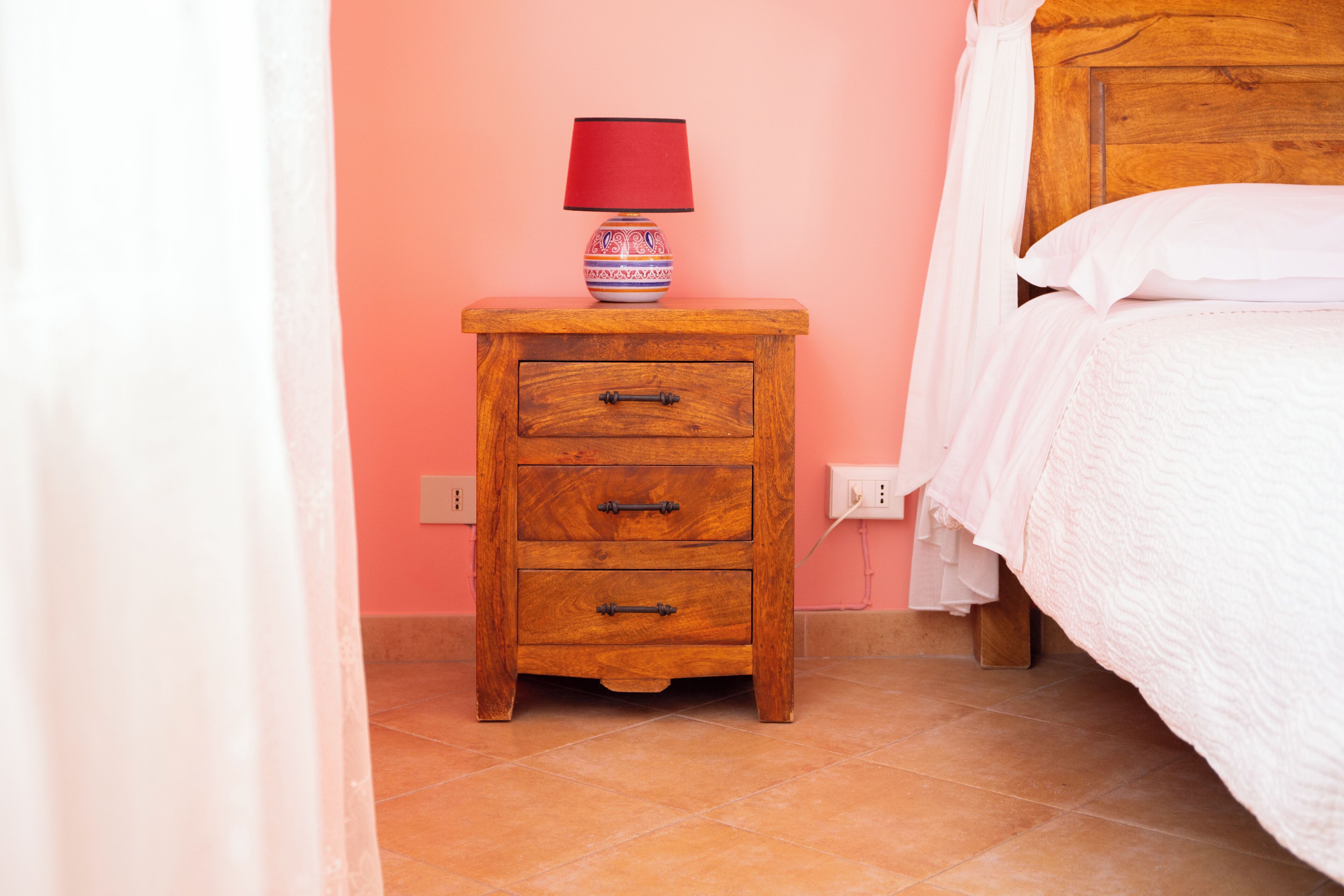 A wooden nightstand with a lamp, a bed with white bedding, and a pink wall.