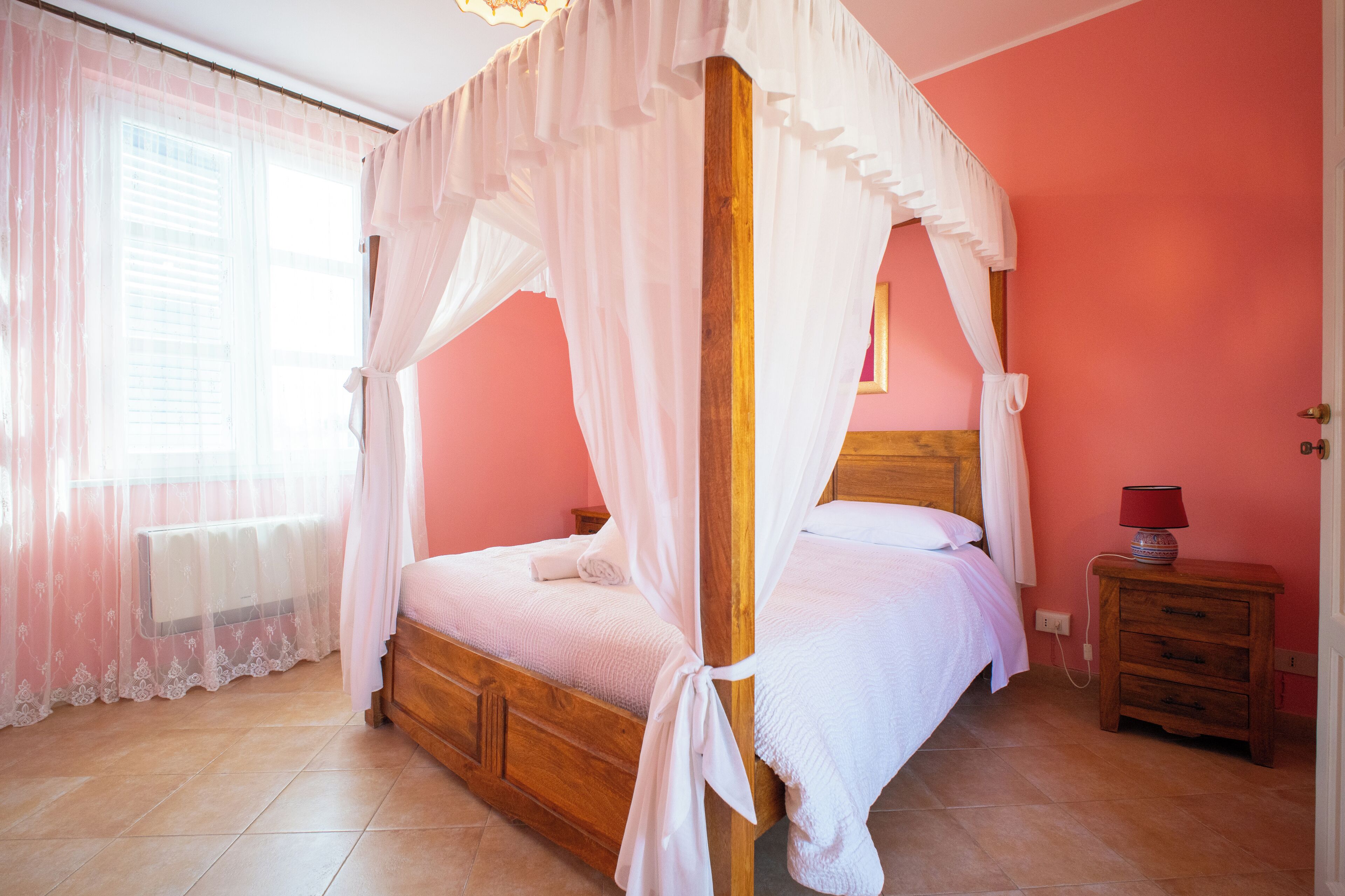 A bedroom with a wooden bed, white bedding, a nightstand with a lamp, and a window with sheer curtains.