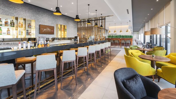 A modern bar area with a long counter, multiple bar stools, and a variety of bottles. - Hilton Garden Inn Windhoek (Windhoek)