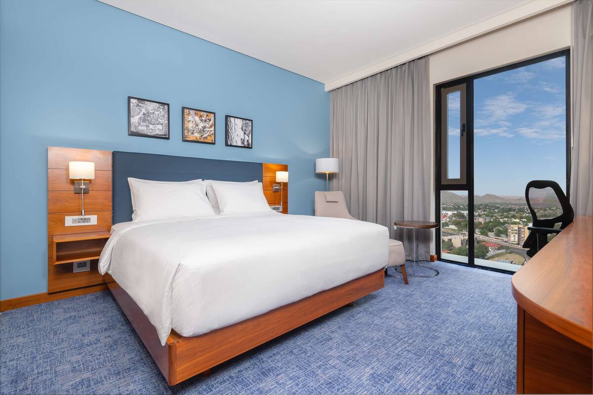 A hotel room with a large bed, two bedside tables with lamps, a desk, and a view of a cityscape through a large window.