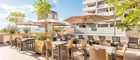 An outdoor patio area with tables and chairs, umbrellas, and a pool in the background.