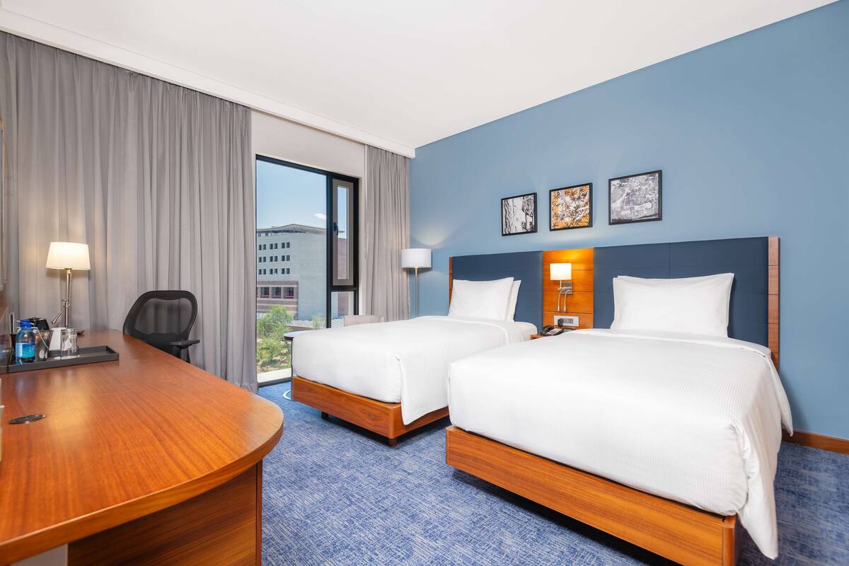 A hotel room with two beds, a desk, and a large window.