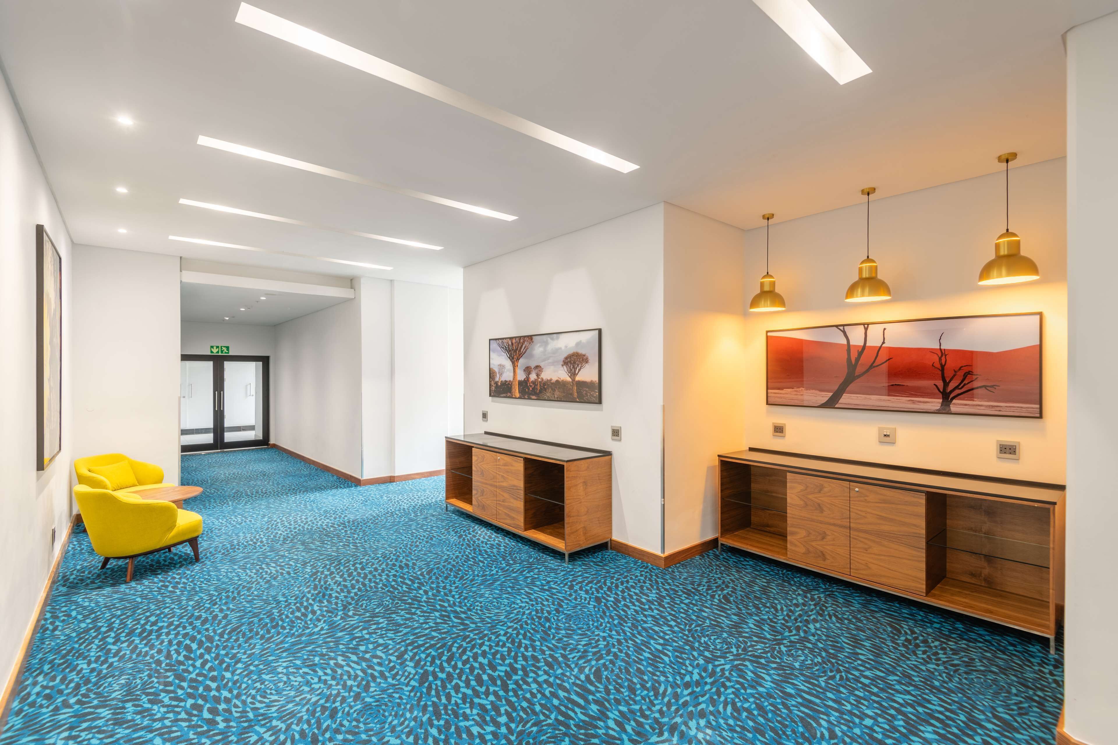 Un vestíbulo de hotel con alfombra azul estampada, mostrador de recepción de madera, sillas amarillas y obras de arte en las paredes.