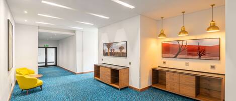 Un vestíbulo de hotel con alfombra azul estampada, mostrador de recepción de madera, sillas amarillas y obras de arte en las paredes.