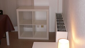 Une chambre avec une table de chevet blanche, une lampe, un radiateur et une fenêtre donnant sur des immeubles.