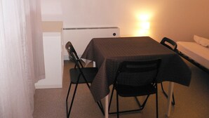 Une petite pièce simple, avec un lit, une table et deux chaises. Un luminaire est fixé au mur au-dessus de la table.