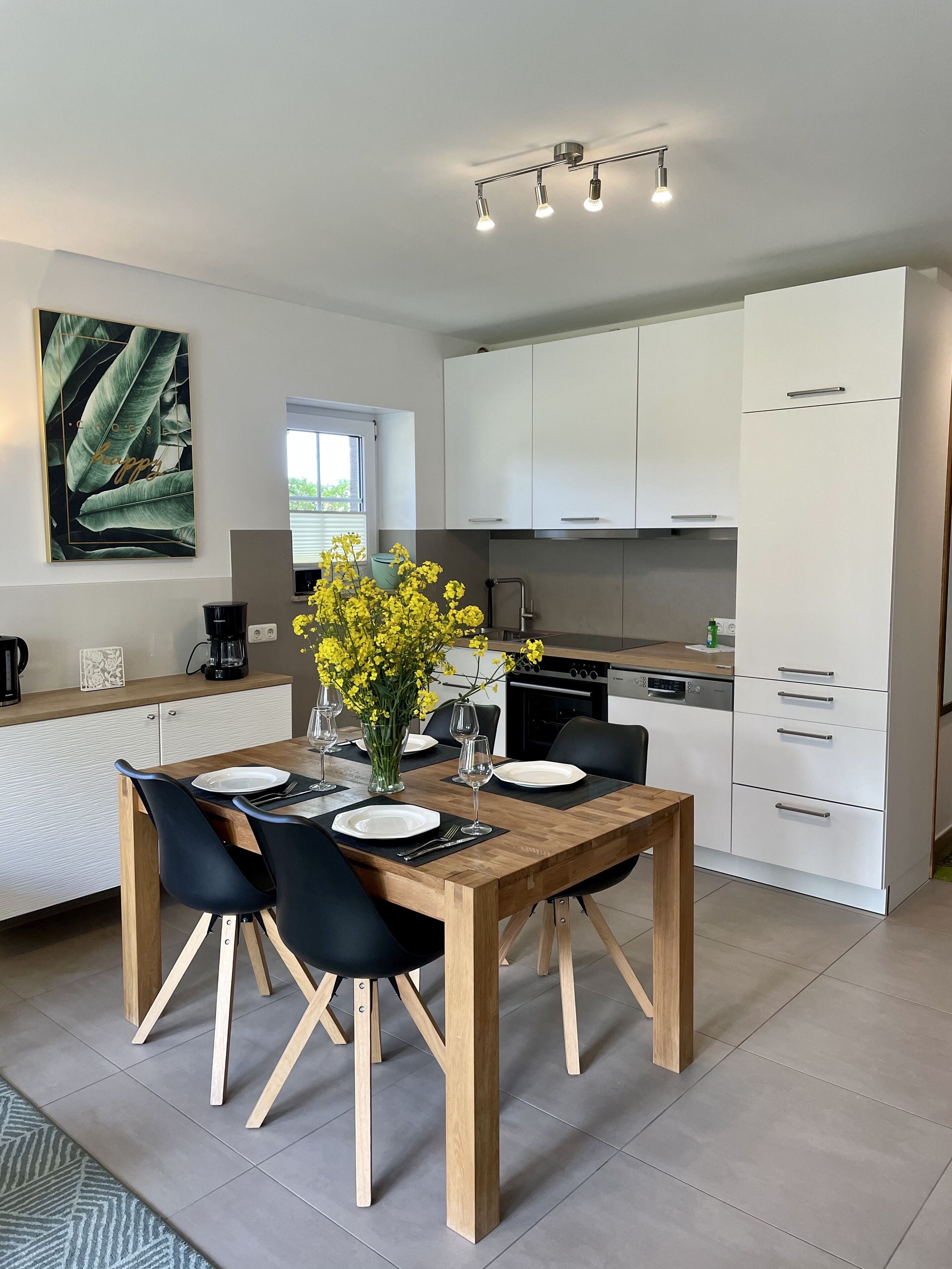 A modern kitchen with white cabinets, a wooden dining table set with chairs, and a vase of yellow flowers.