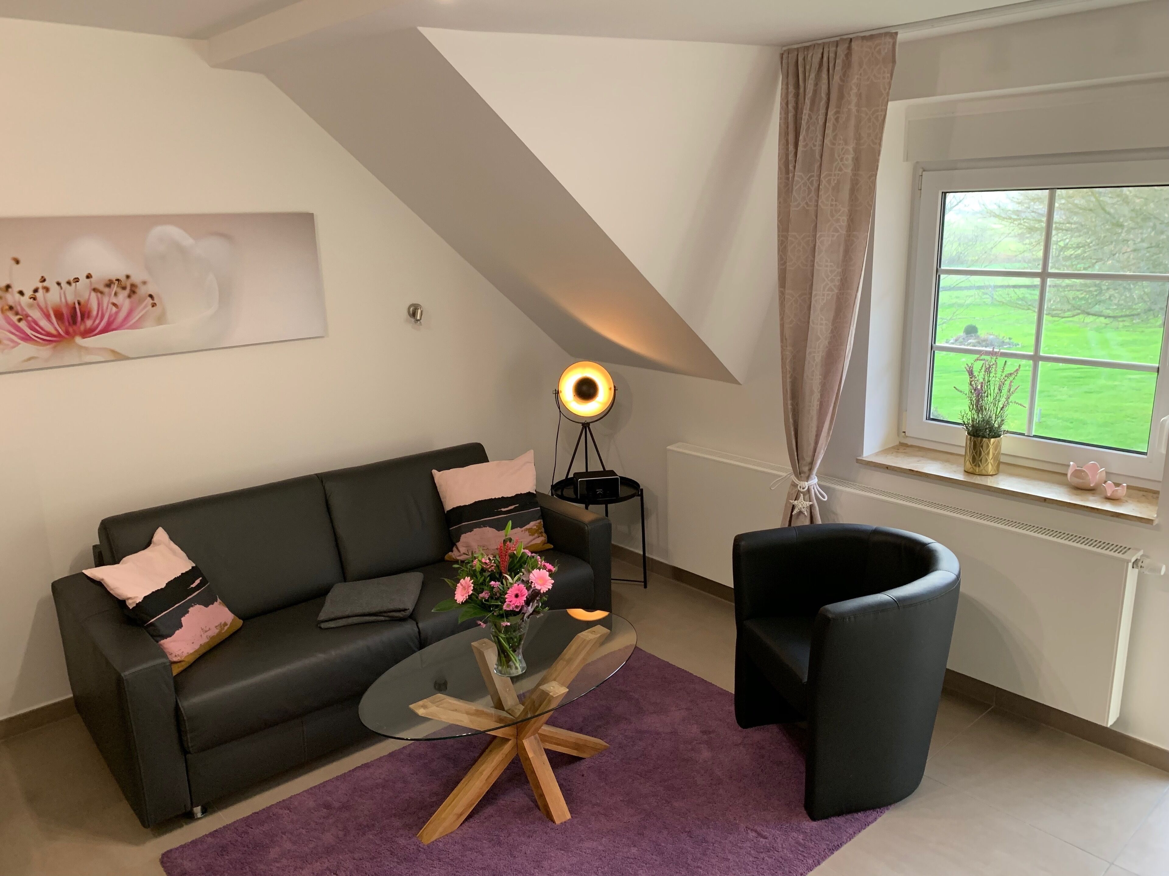 Un salon moderne avec un canapé noir, une table basse en verre, un lampadaire et une vue sur un jardin verdoyant par la fenêtre.