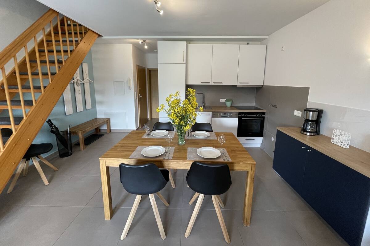 A modern kitchen with white cabinets, a wooden dining table, two black chairs, and a vase of yellow flowers.