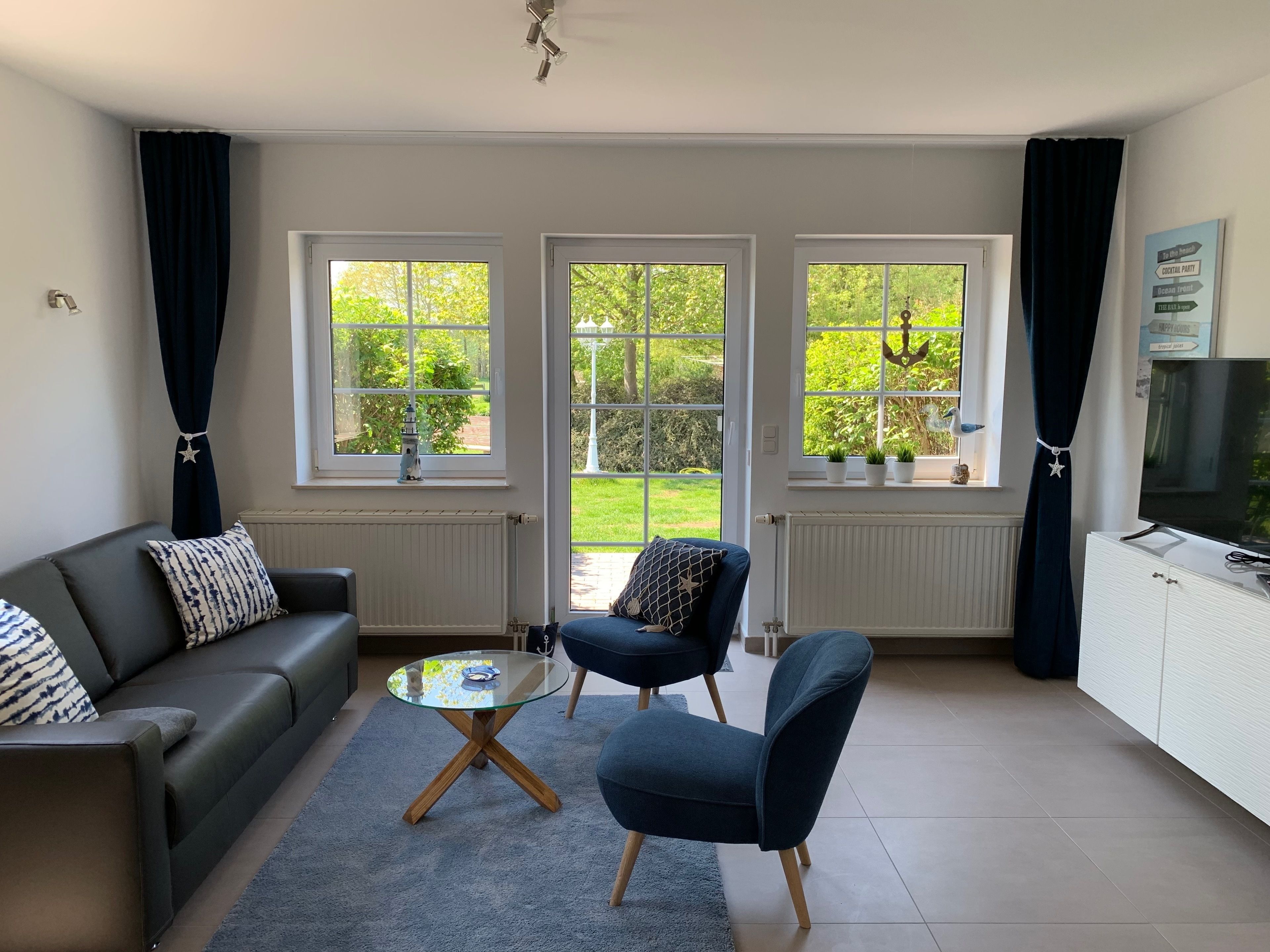 A modern living room with a sofa, two armchairs, a glass coffee table, and a TV on a cabinet. Large windows offer a view of a garden.