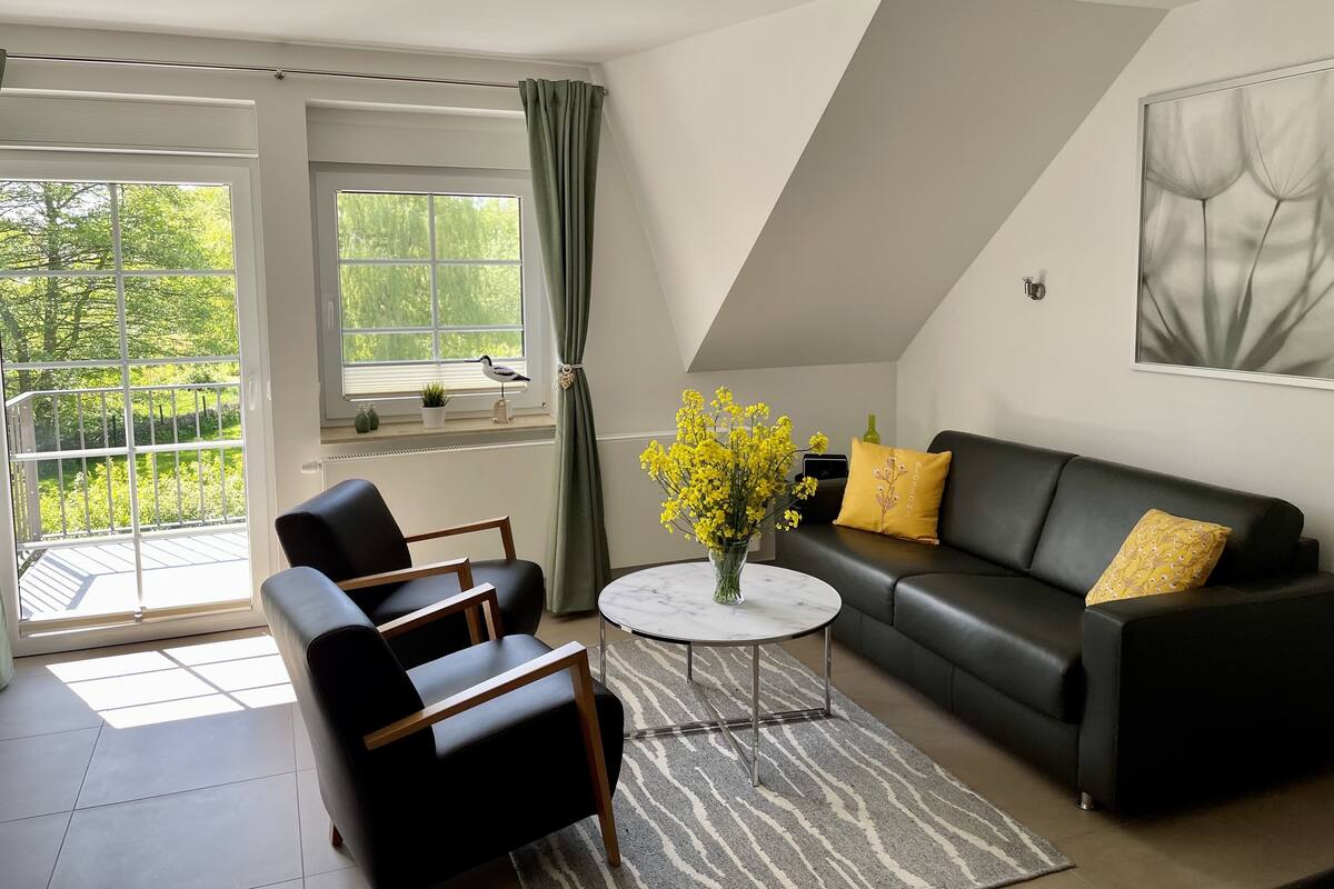 A modern living room with a black leather sofa, a black armchair, and a round coffee table. There is a large window with a view of greenery outside.