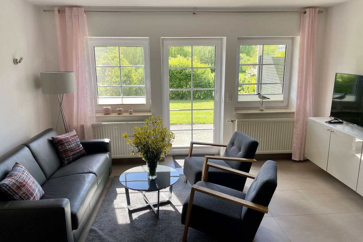 A modern living room with a black leather sofa, a grey armchair, a round glass coffee table, and a TV on a white cabinet. Large windows with pink curtains offer views of a garden.