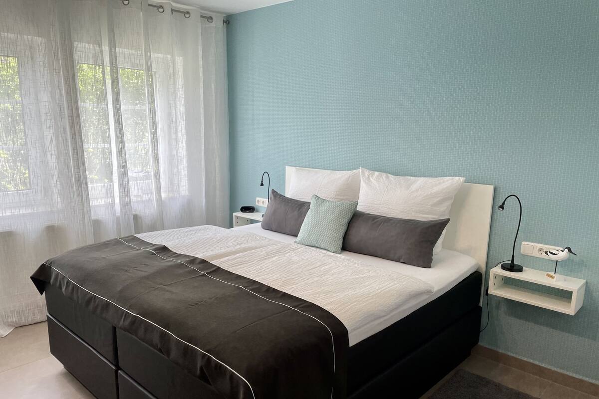 A modern bedroom with a large bed, bedside tables, a nightstand, and a window with sheer curtains.