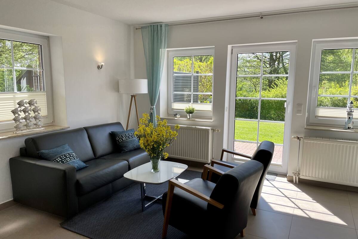 A modern living room with a black sofa, a coffee table, and a chair. There are large windows with curtains, a radiator, and a small plant on the windowsill.
